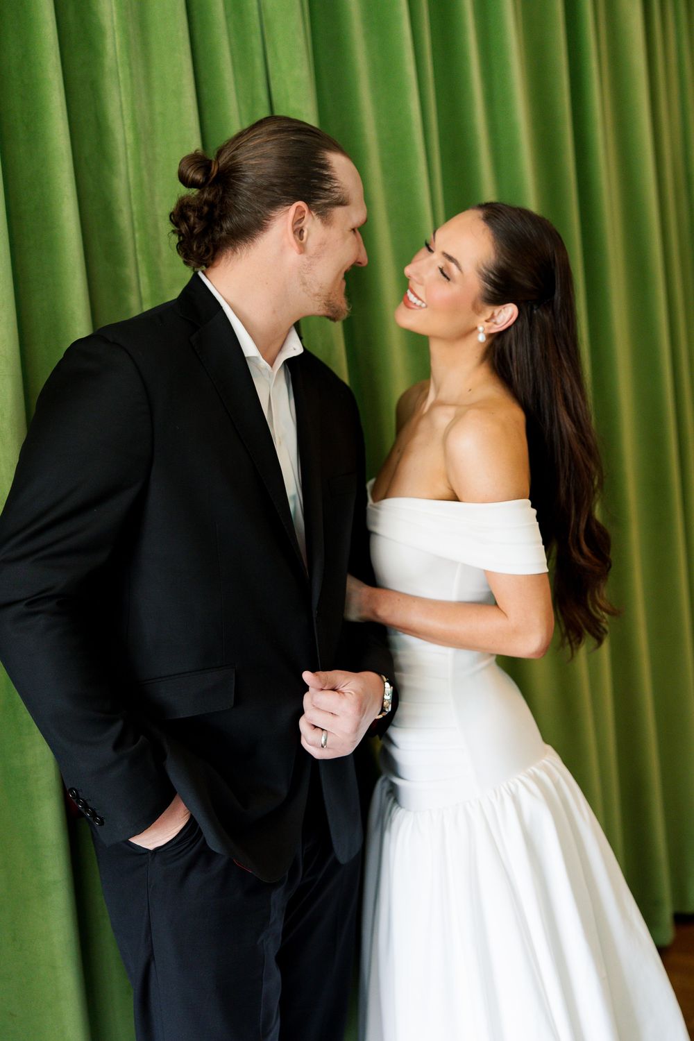 A bride in a white off-shoulder gown and groom in a black suit share a romantic moment before green velvet curtains.
