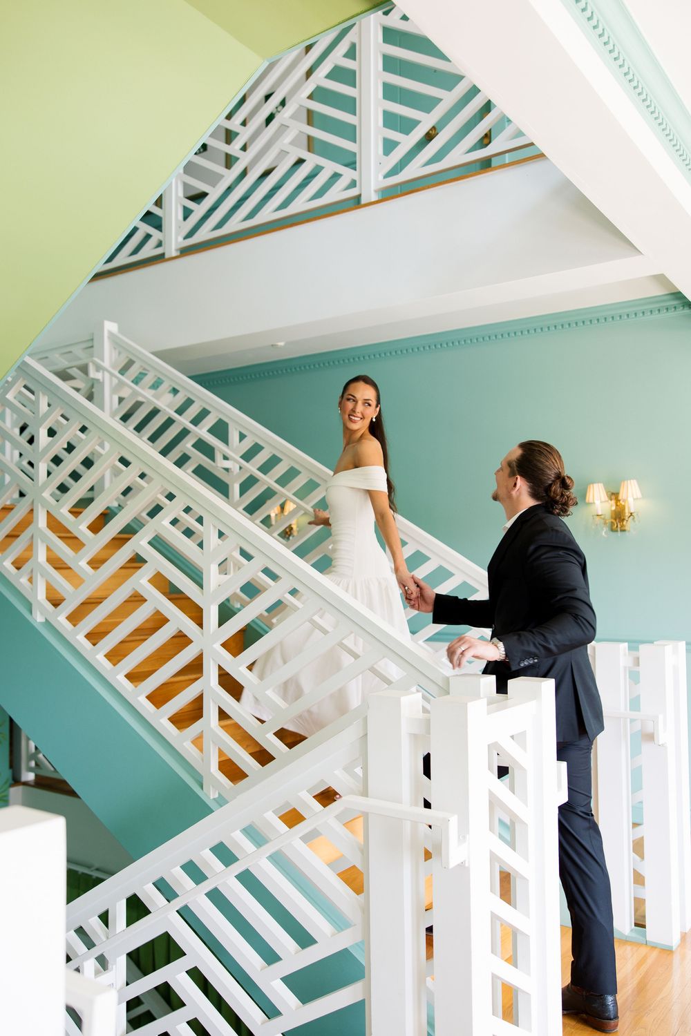Bride and groom holding hands on a white geometric staircase with teal walls in an elegant venue.