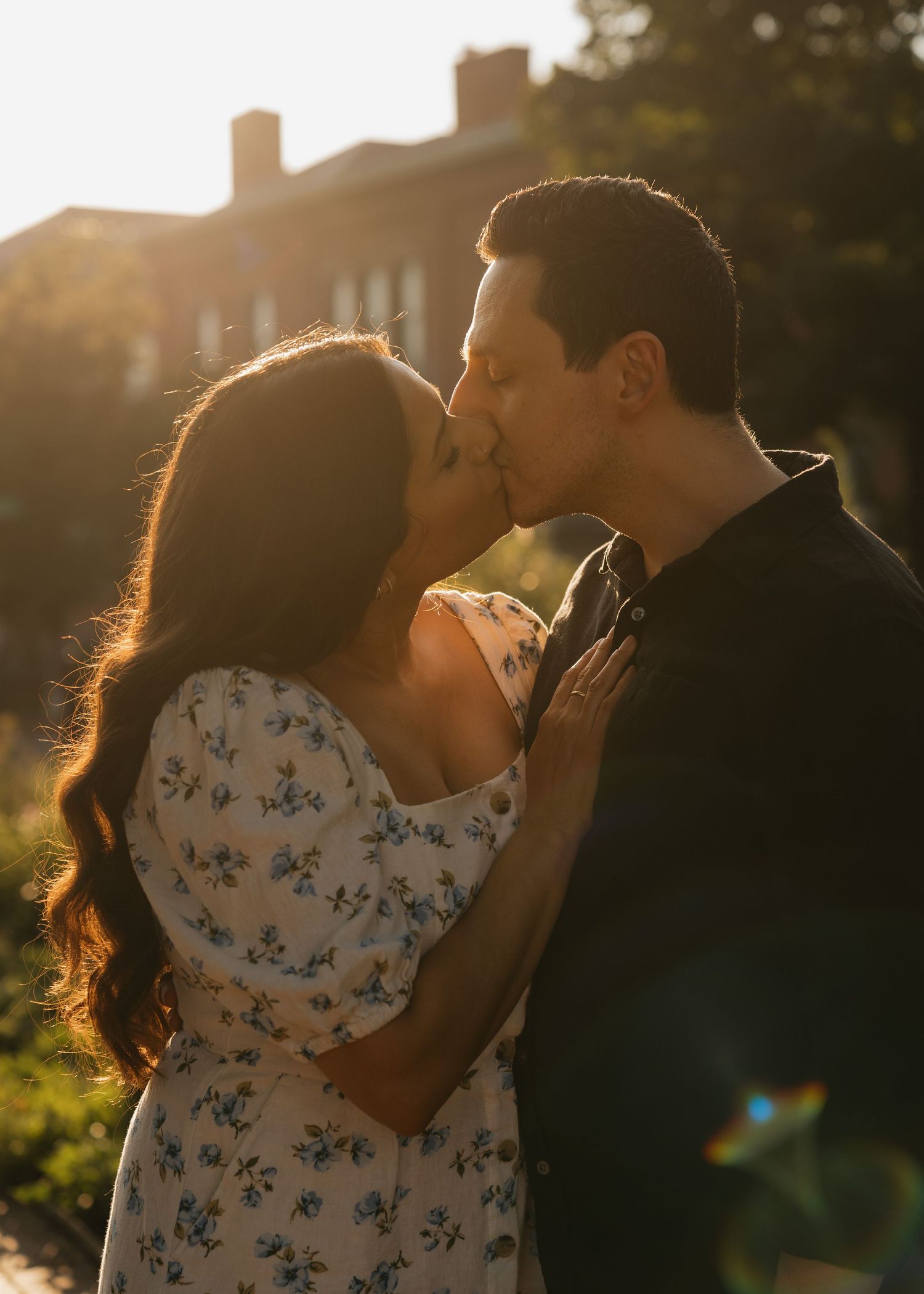 Romantic couple in floral dress and dark suit embraces and kisses in warm sunset lighting.