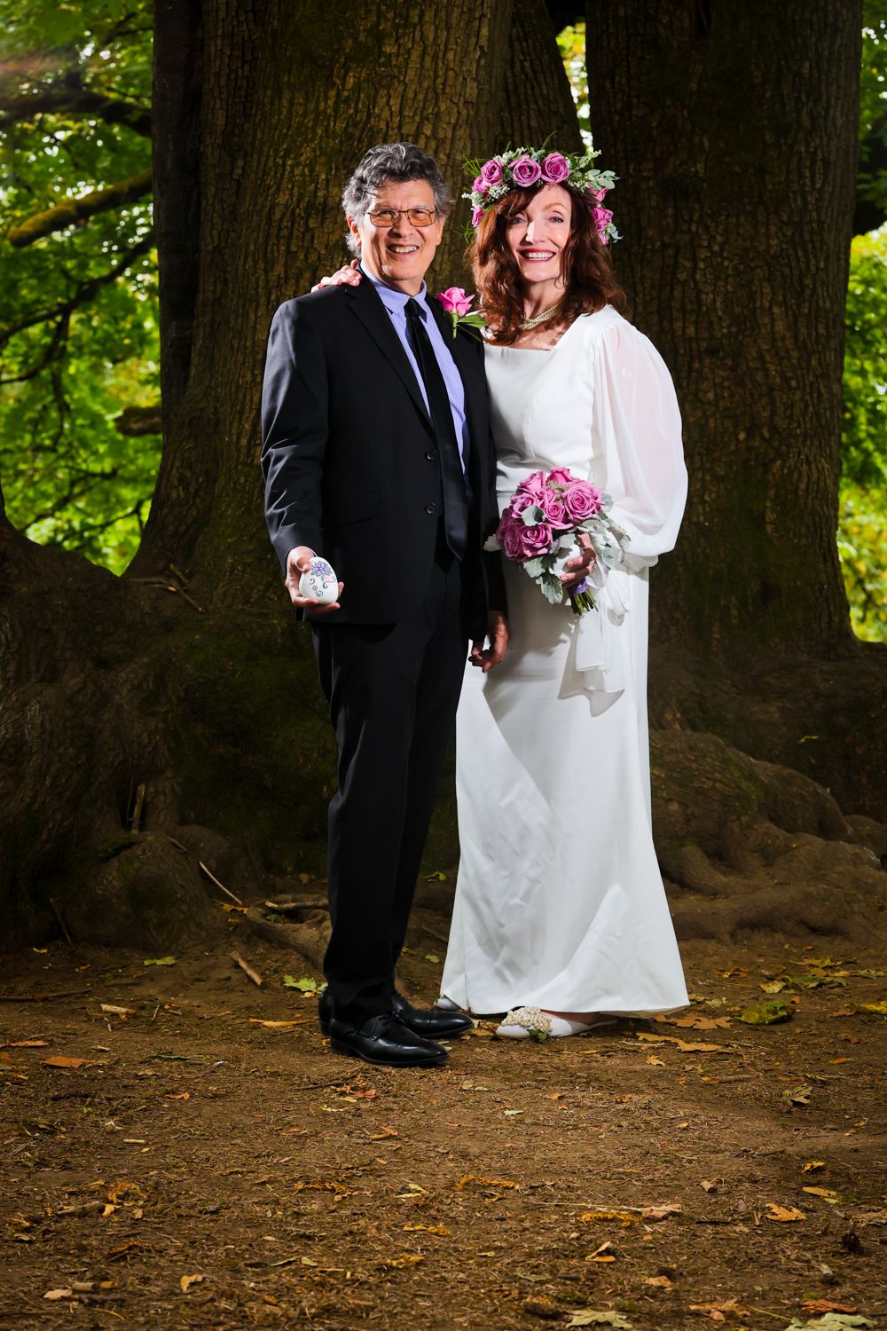 A couple poses for a wedding photo in front of large tree trunks, with the bride wearing a floral crown and white dress.