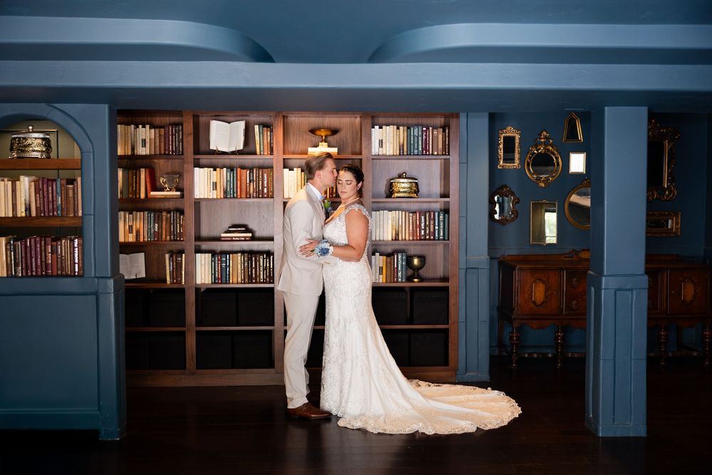 A wedding couple embraces in front of built-in bookshelves in a beautifully lit library setting.