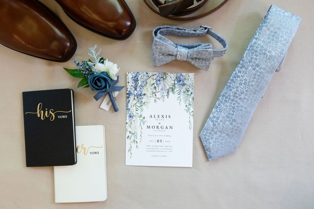 Wedding details flat lay with brown dress shoes, grey tie, invitation card, vows books and floral decor on light background.