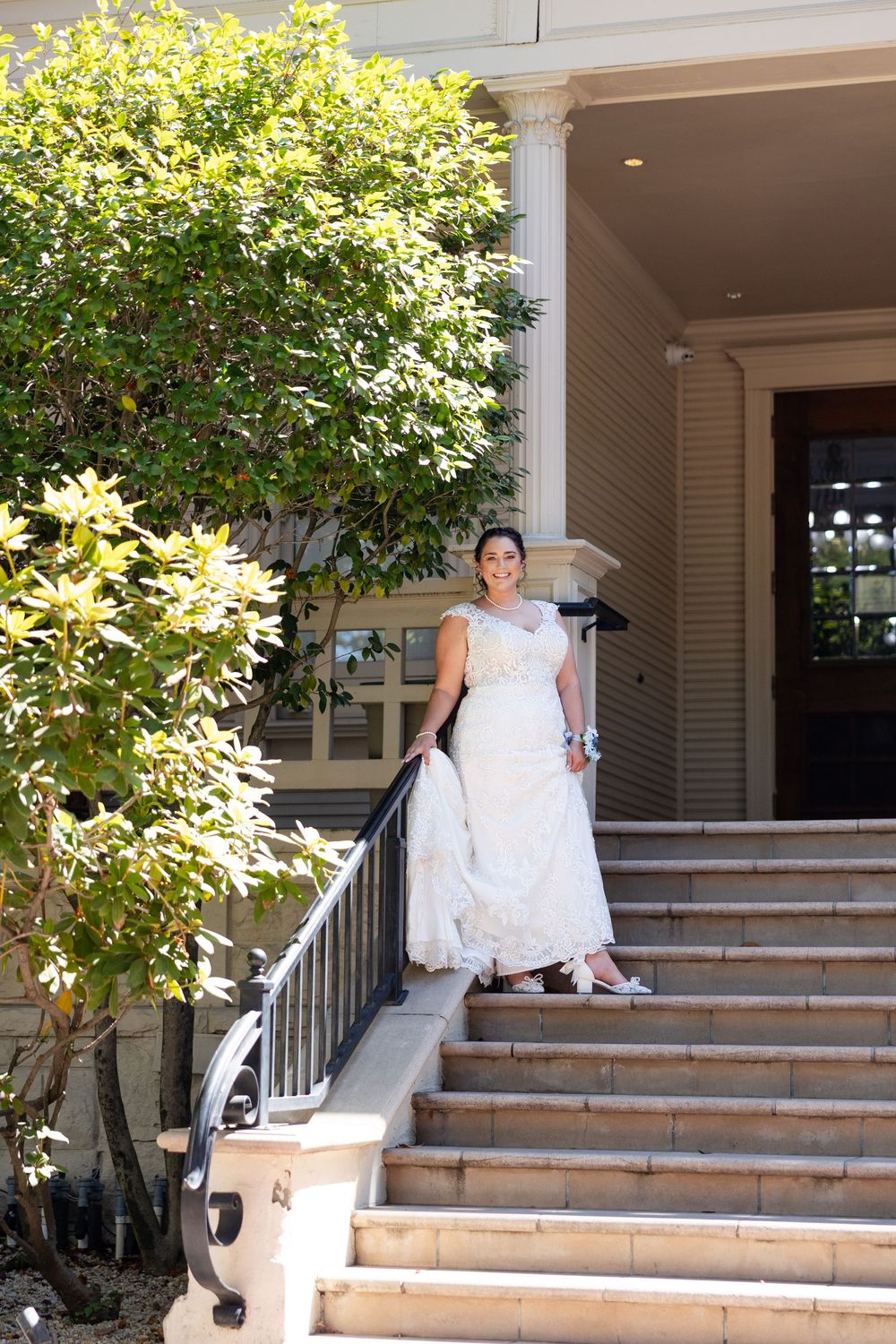 A person in an elegant white wedding dress stands on stone steps outside a building entrance framed by lush greenery.