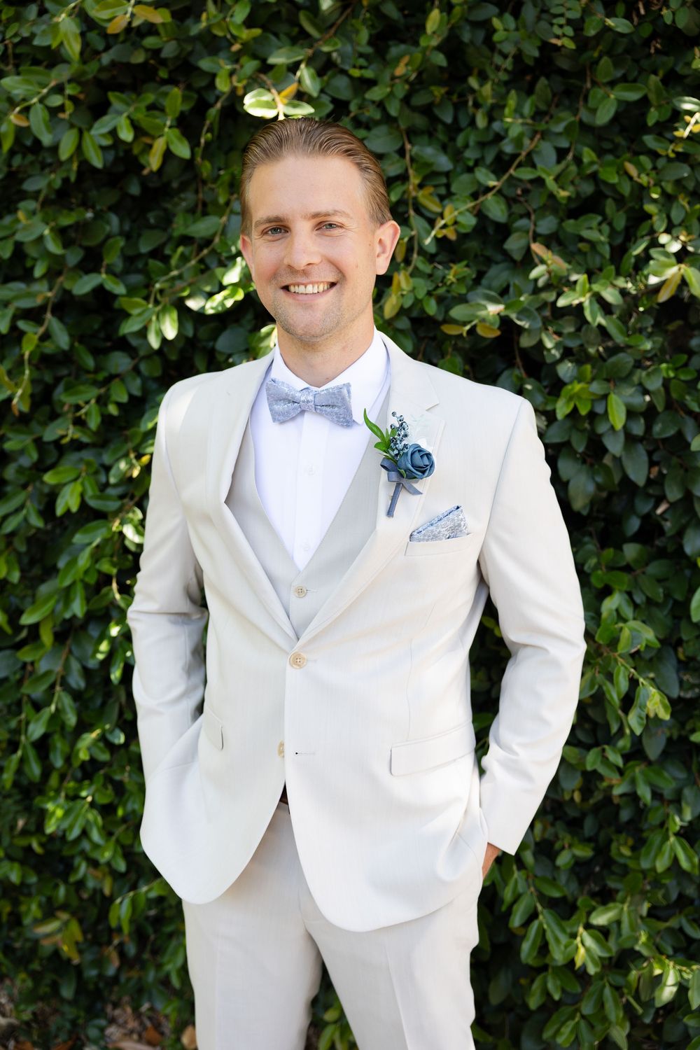 A groom wearing a white suit and bow tie poses in front of a green hedge wall for wedding photos.