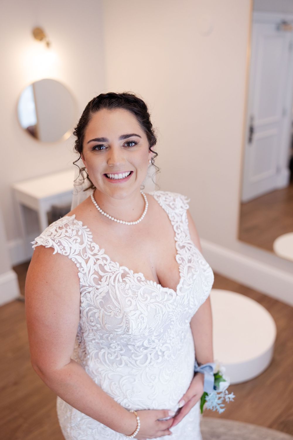A bride wearing a beautiful lace wedding dress with pearl necklace poses in a bridal suite.