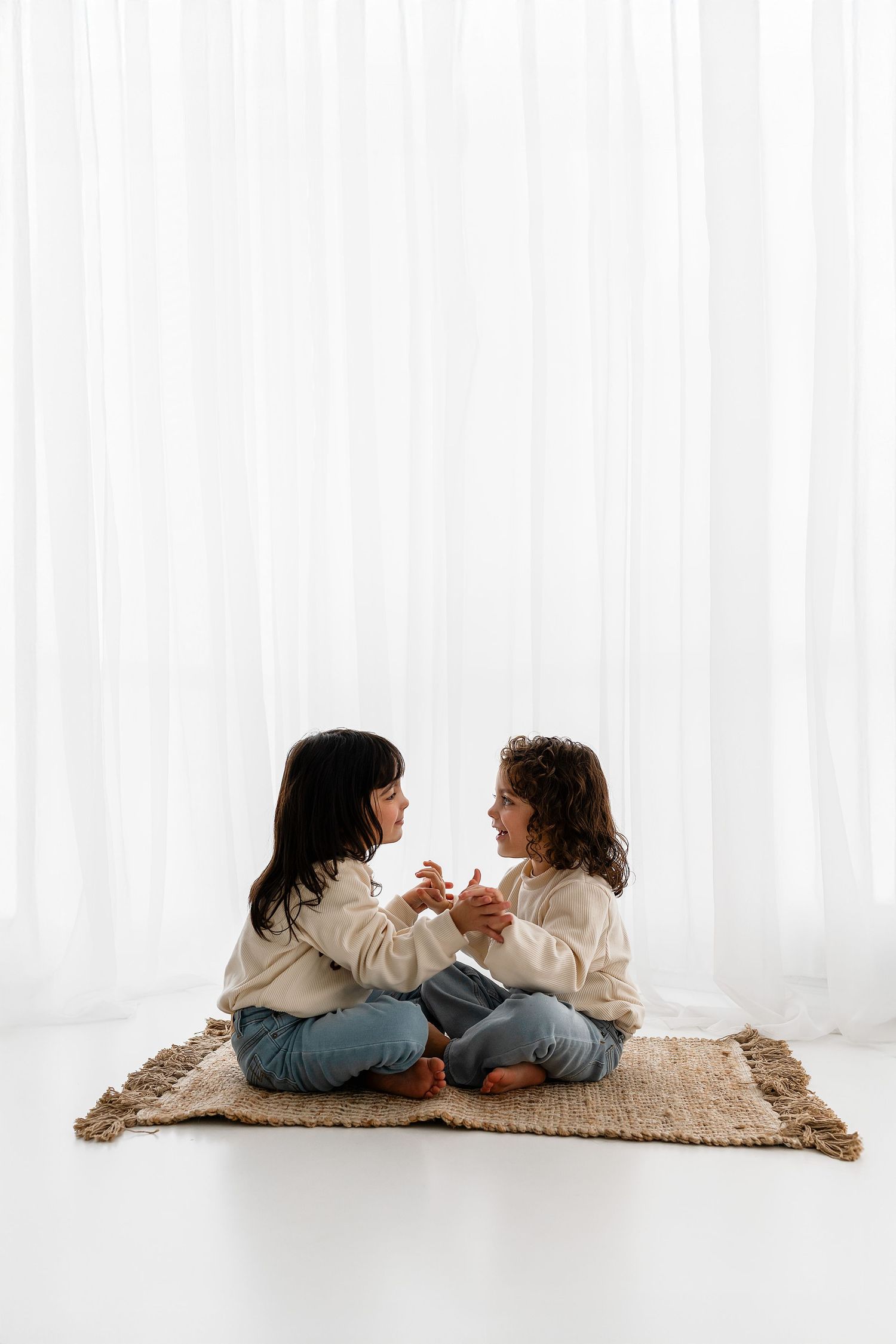 Two children in matching cream sweaters sitting cross-legged on a woven mat, sharing a playful moment together.