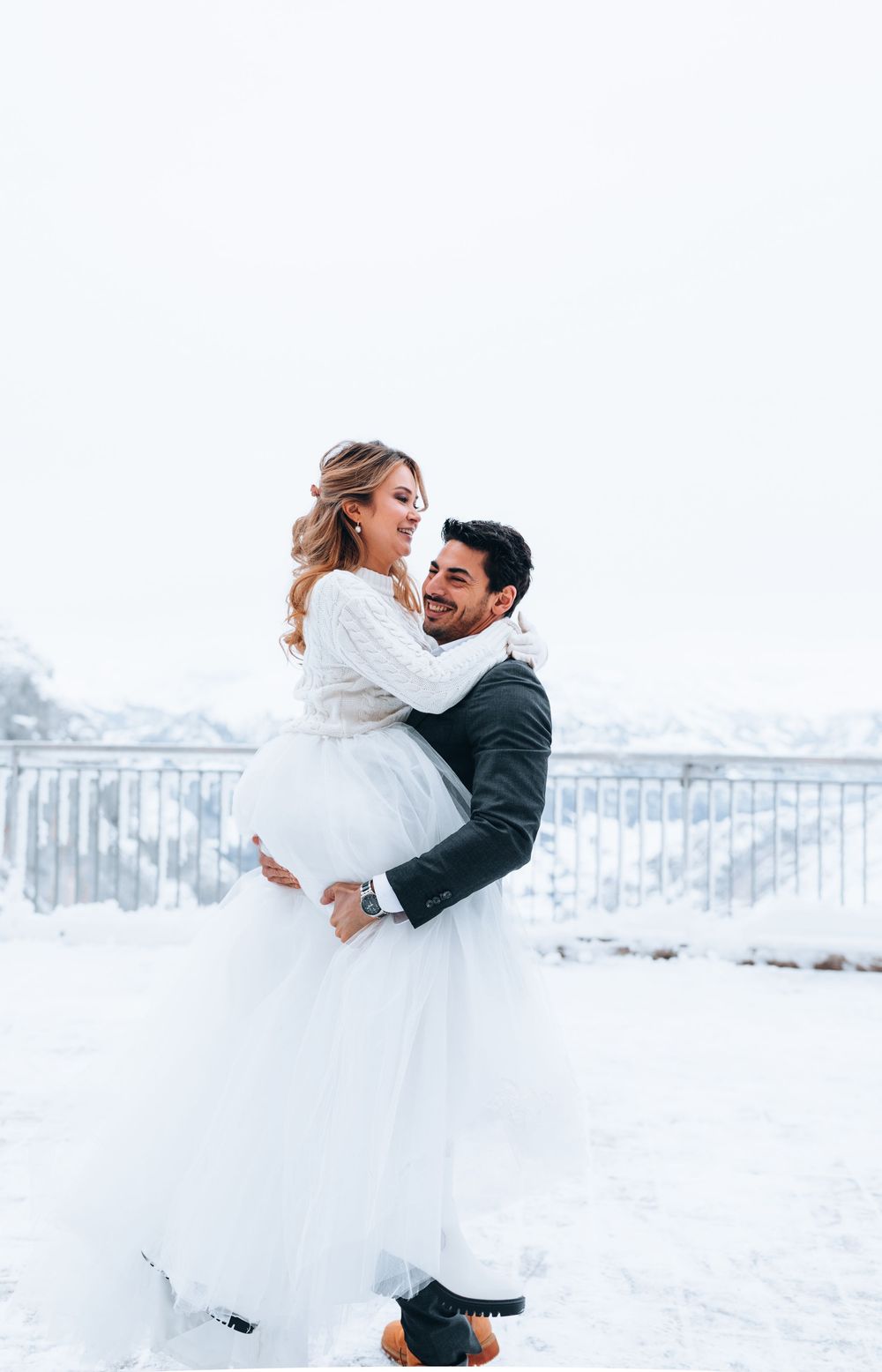 A couple embraces in a romantic winter scene with snow-covered mountains and railing in the background.
