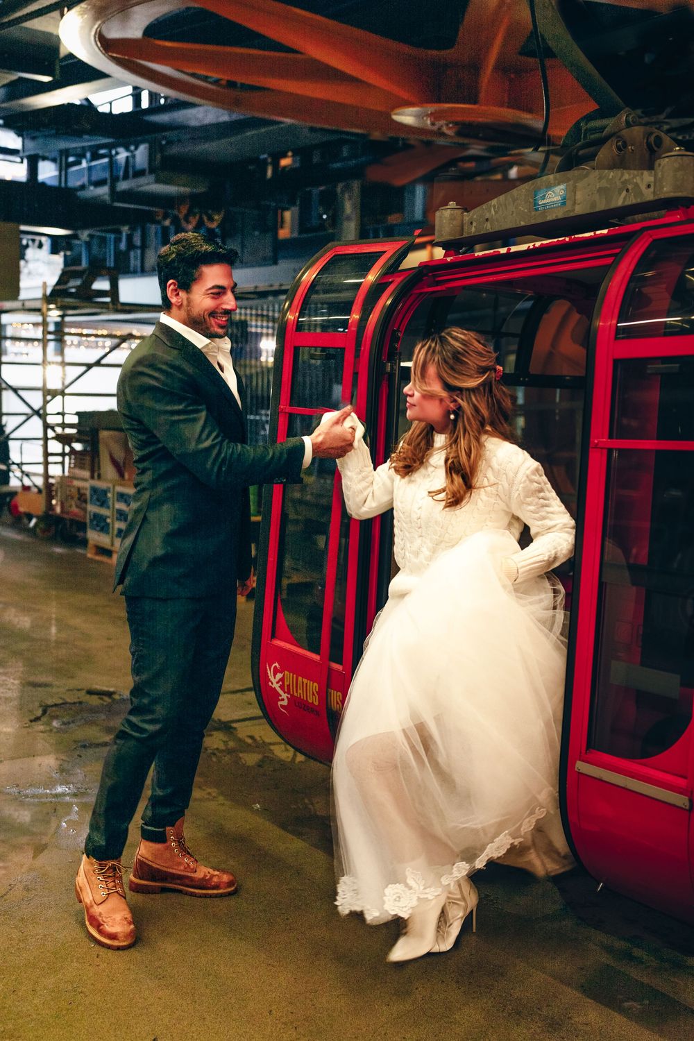 A couple in wedding attire hold hands while walking through an industrial area with red train cars in the background.