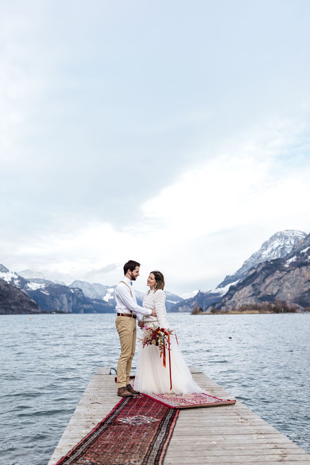 A couple shares a romantic moment on a wooden dock overlooking a scenic mountain lake with snow-capped peaks in the background.