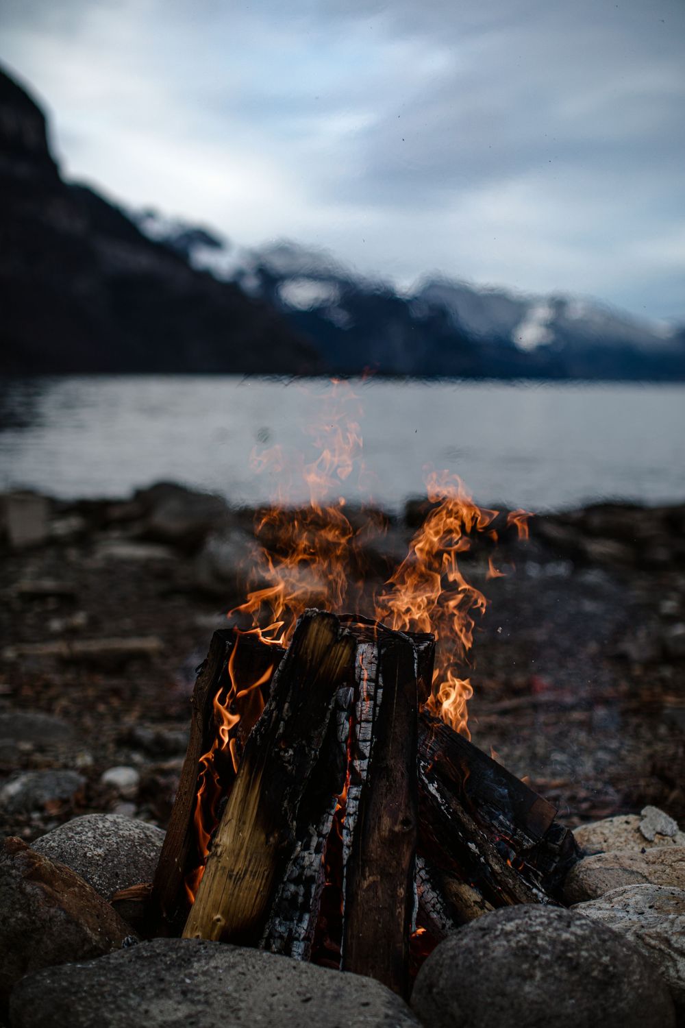 Glowing campfire burning brightly on rocky shore of misty mountain lake with dramatic cloudy skies in background.