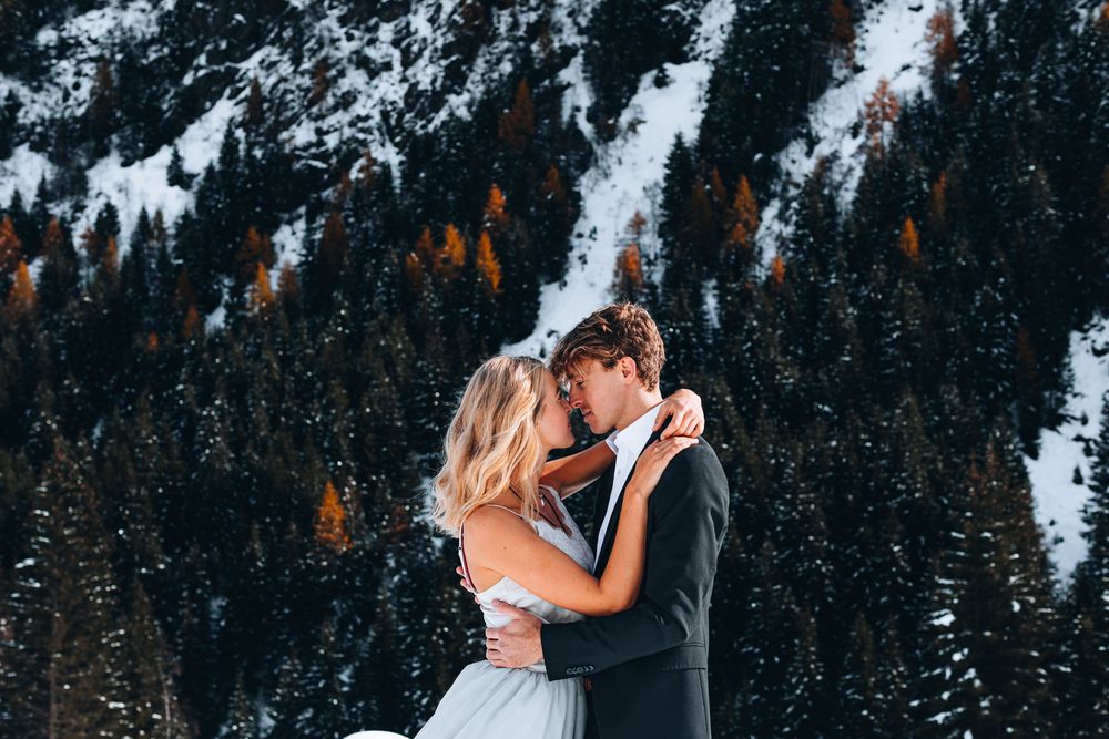 Couple in wedding attire embrace against snowy mountainscape with evergreen trees and autumn foliage in background.