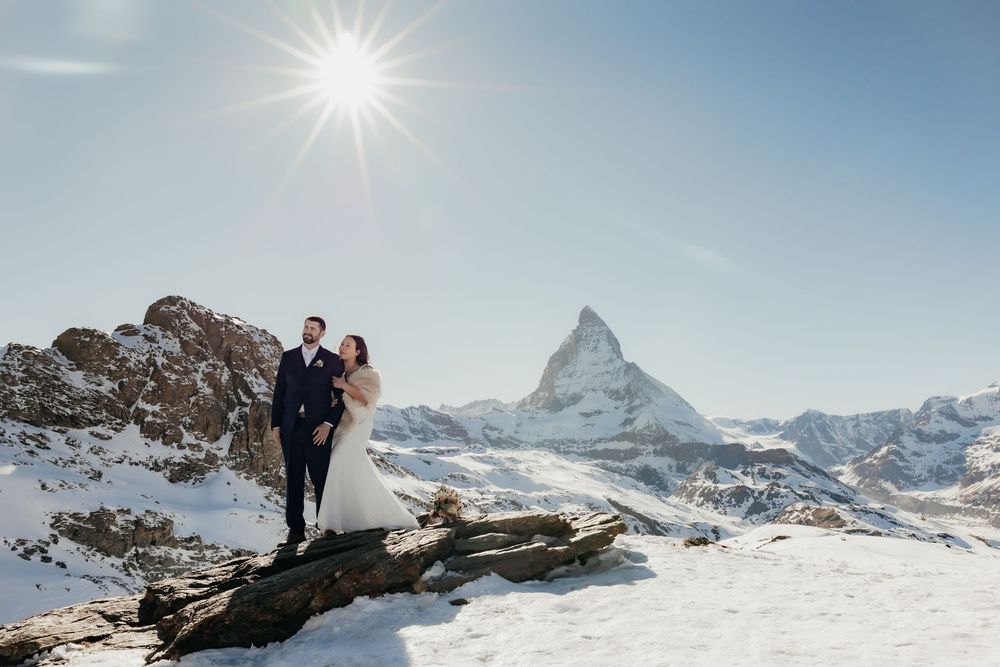 Couple stands on snowy mountain peak with majestic Matterhorn mountain visible in background on sunny winter day.
