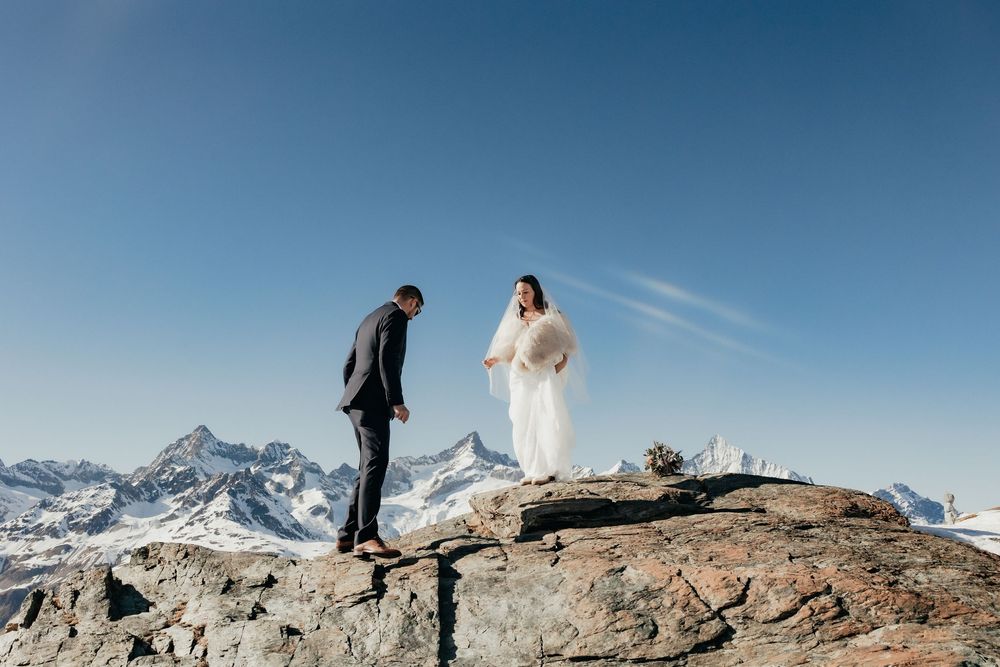 A couple stands atop a rocky mountain peak with snow-capped mountains in the background under a bright blue sky.