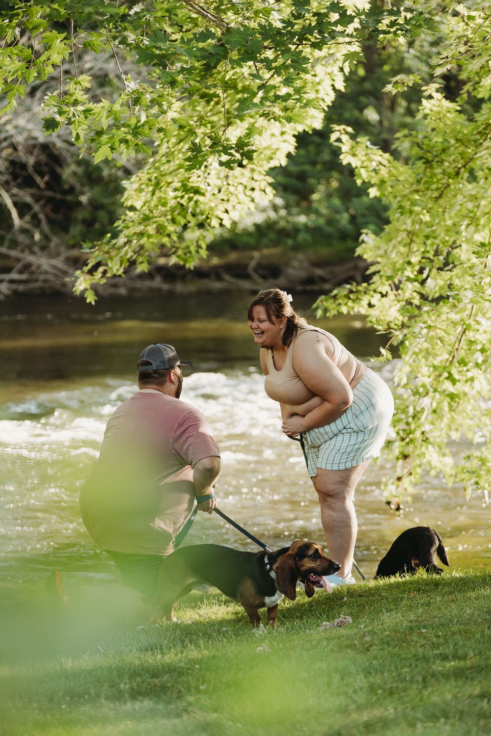 A couple and their dog enjoy a moment together by a flowing river on a sunny afternoon under leafy tree branches.