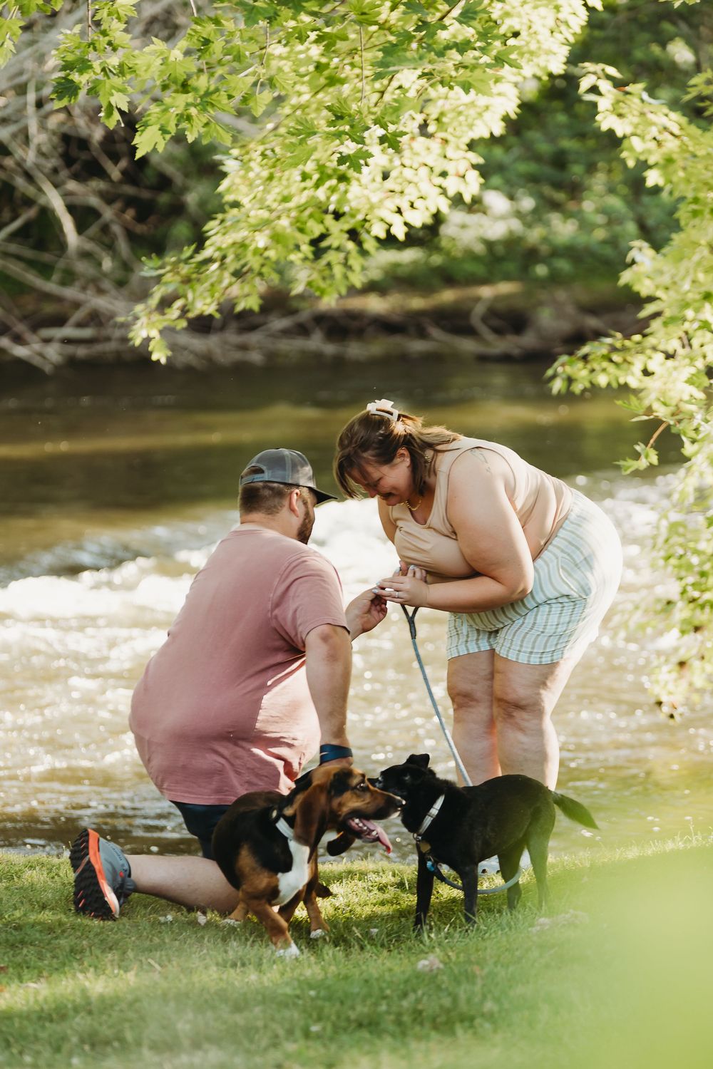 Two people and a dog share a tender moment by a peaceful riverside surrounded by lush green trees and flowing water.