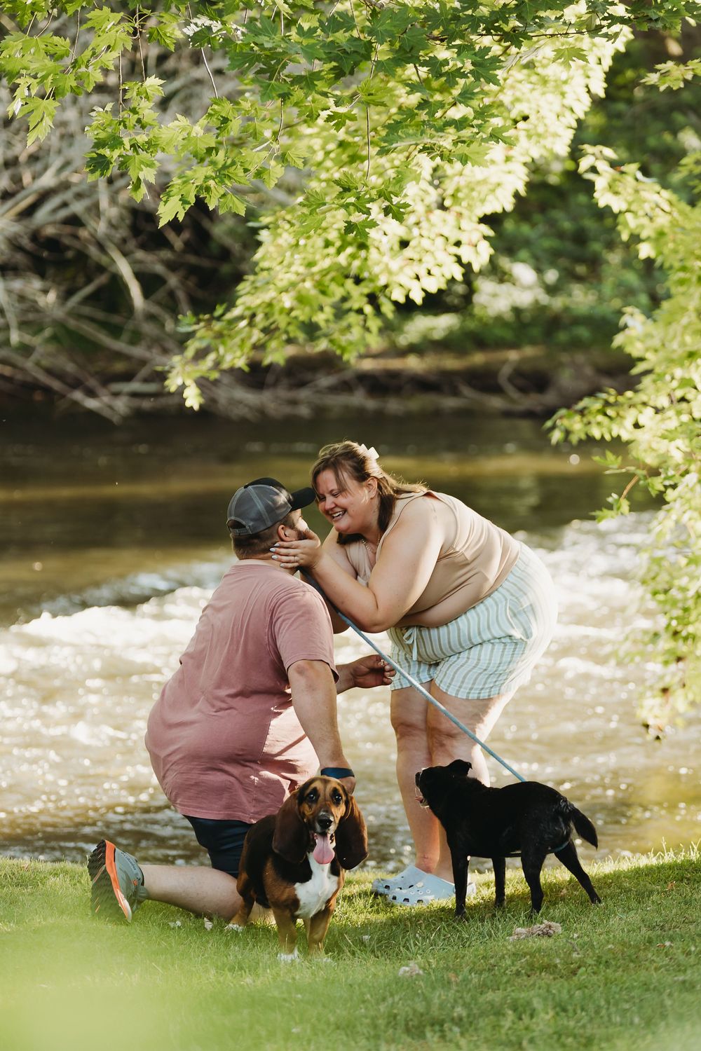 A couple and their black dog interact lovingly near a flowing river on a sunny day amidst green foliage.