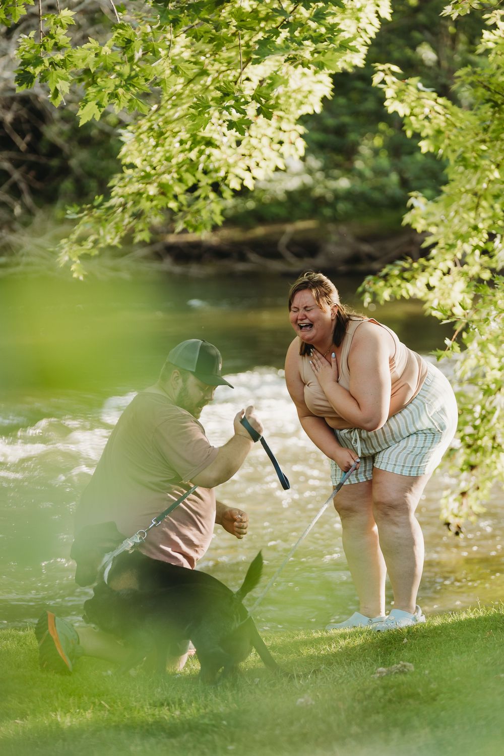 Sweet proposal moment captured by the water's edge under overhanging tree branches on a sunny day.
