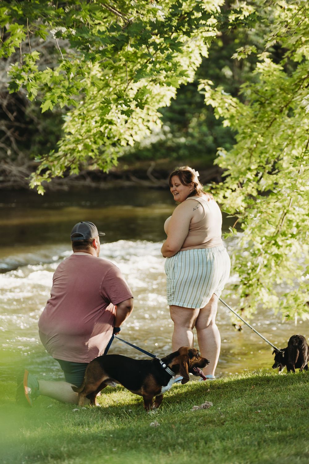 Two people sit together on grassy riverbank under dappled sunlight enjoying a peaceful moment by flowing water.