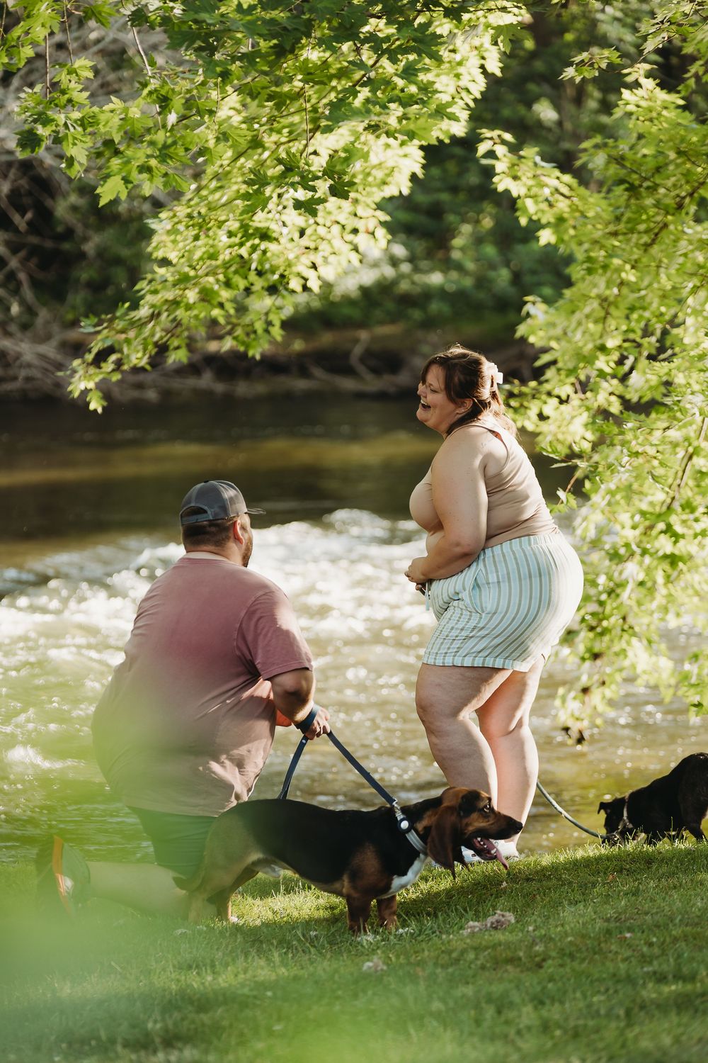 Two people enjoying a relaxing afternoon by a river with their small dog beneath green overhanging tree branches.