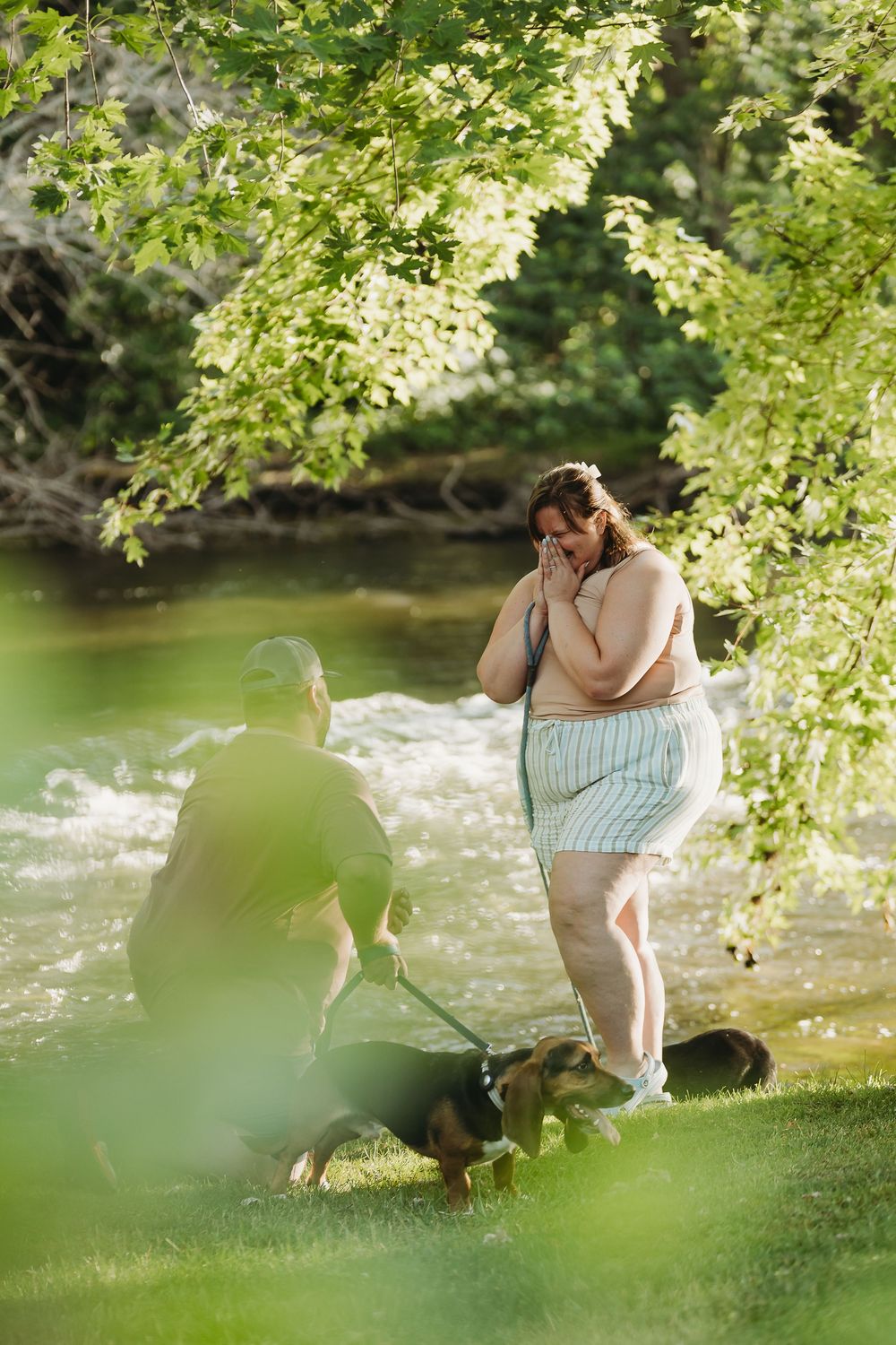 Two people and a dog spend a peaceful afternoon by a serene river surrounded by overhanging green trees.