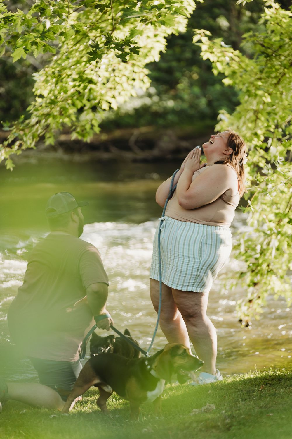 A person enjoys a peaceful moment by a flowing river on a sunny day under green overhanging tree branches.