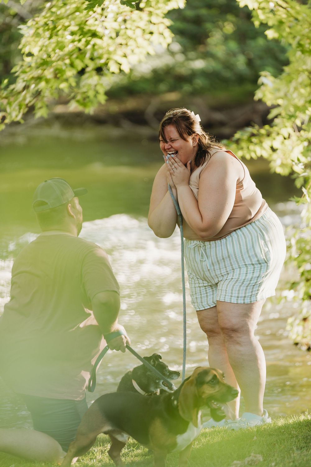 An emotional outdoor moment captured by a creek under flowering tree branches on a sunny summer day.
