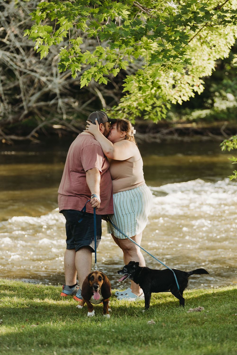 A couple shares an intimate moment by a river while their two dogs, a beagle and black lab, stand nearby.