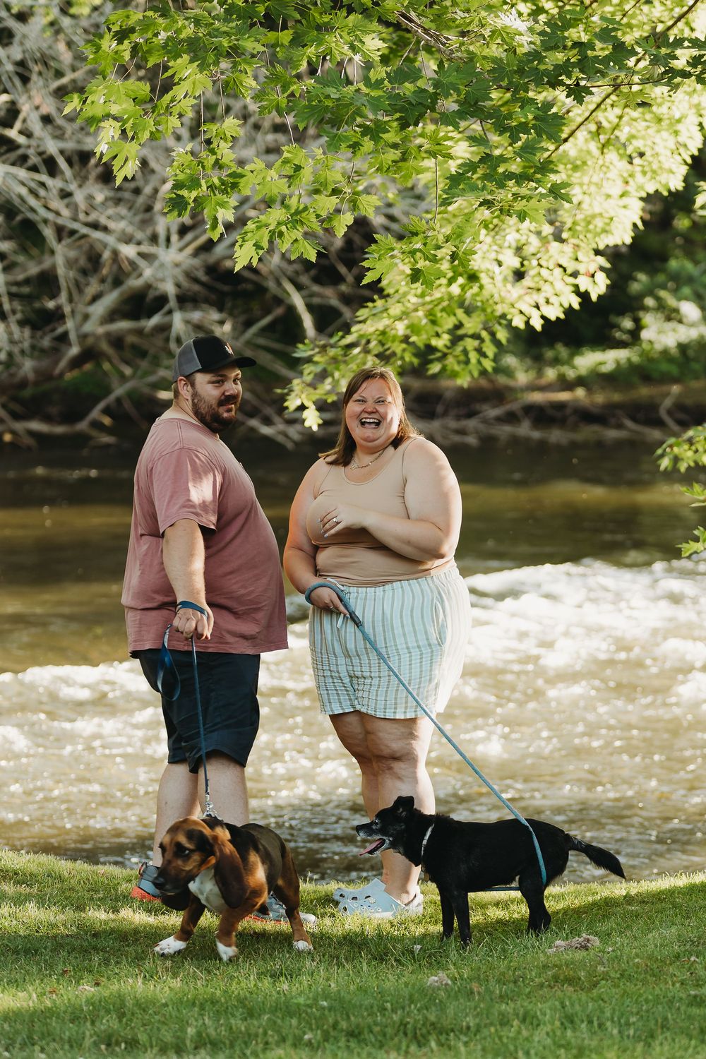 Two people walking their Basset Hound and black Labrador dogs along a riverbank on a sunny spring day.