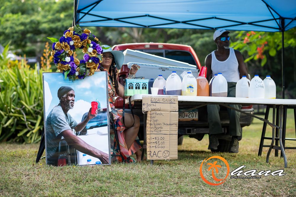 An outdoor vendor stand with colorful flowers and drinks set up under a blue canopy tent on a grassy lawn.