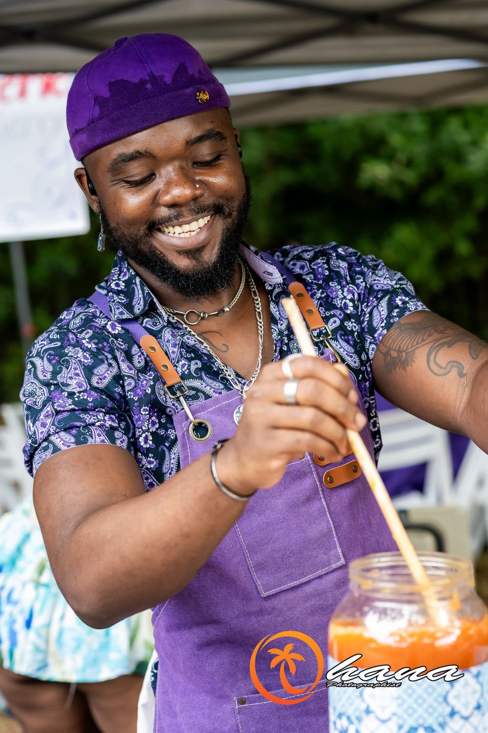 A chef in a purple bandana and apron stirs a drink while smiling during a culinary event outdoors.