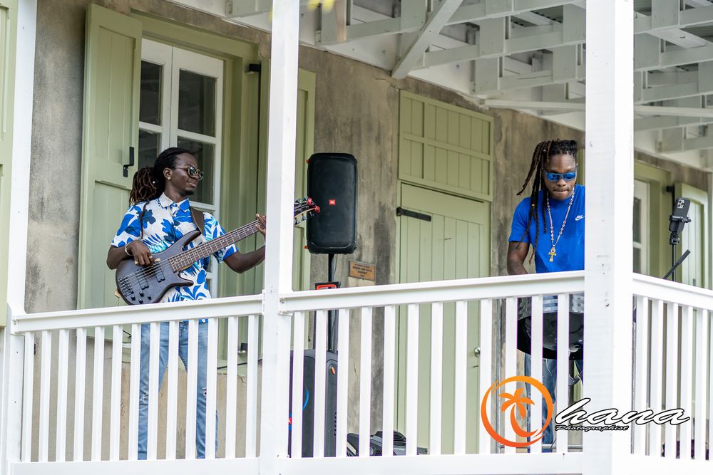 Two musicians perform on a white wooden porch with a guitar and sound equipment.