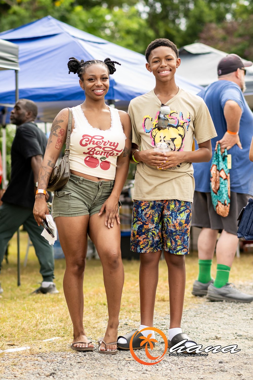 Two people standing together at an outdoor event with pop-up tents, wearing casual summer clothing and smiling.
