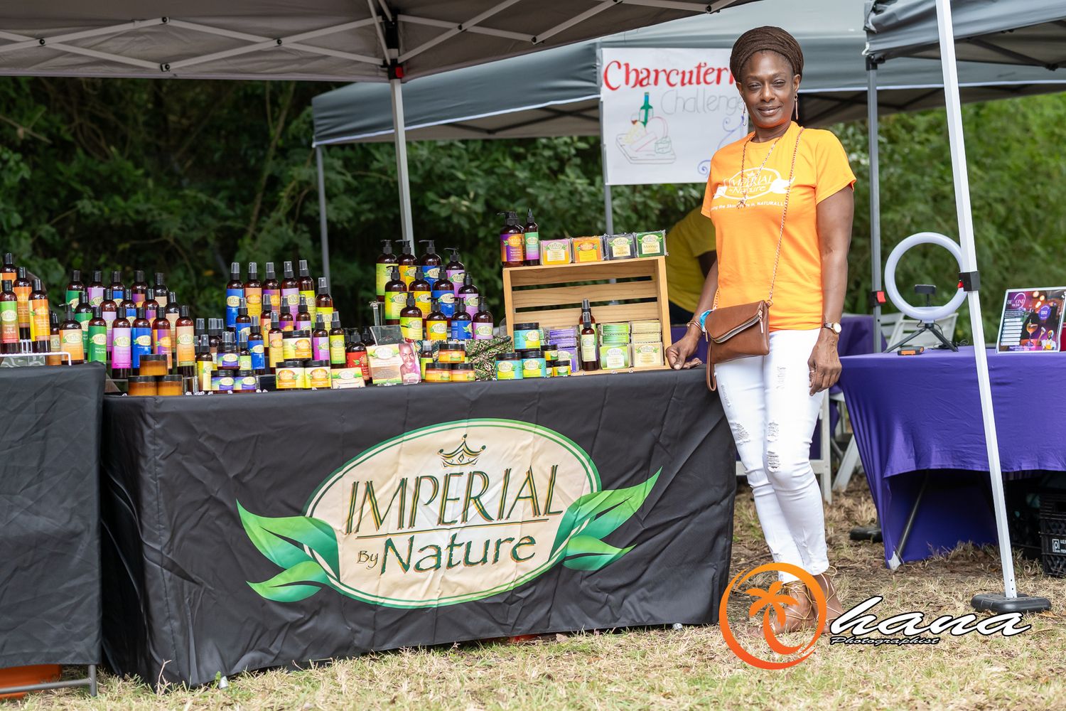 Small business vendor stands at outdoor market booth displaying Imperial Nature products under white canopy tent.