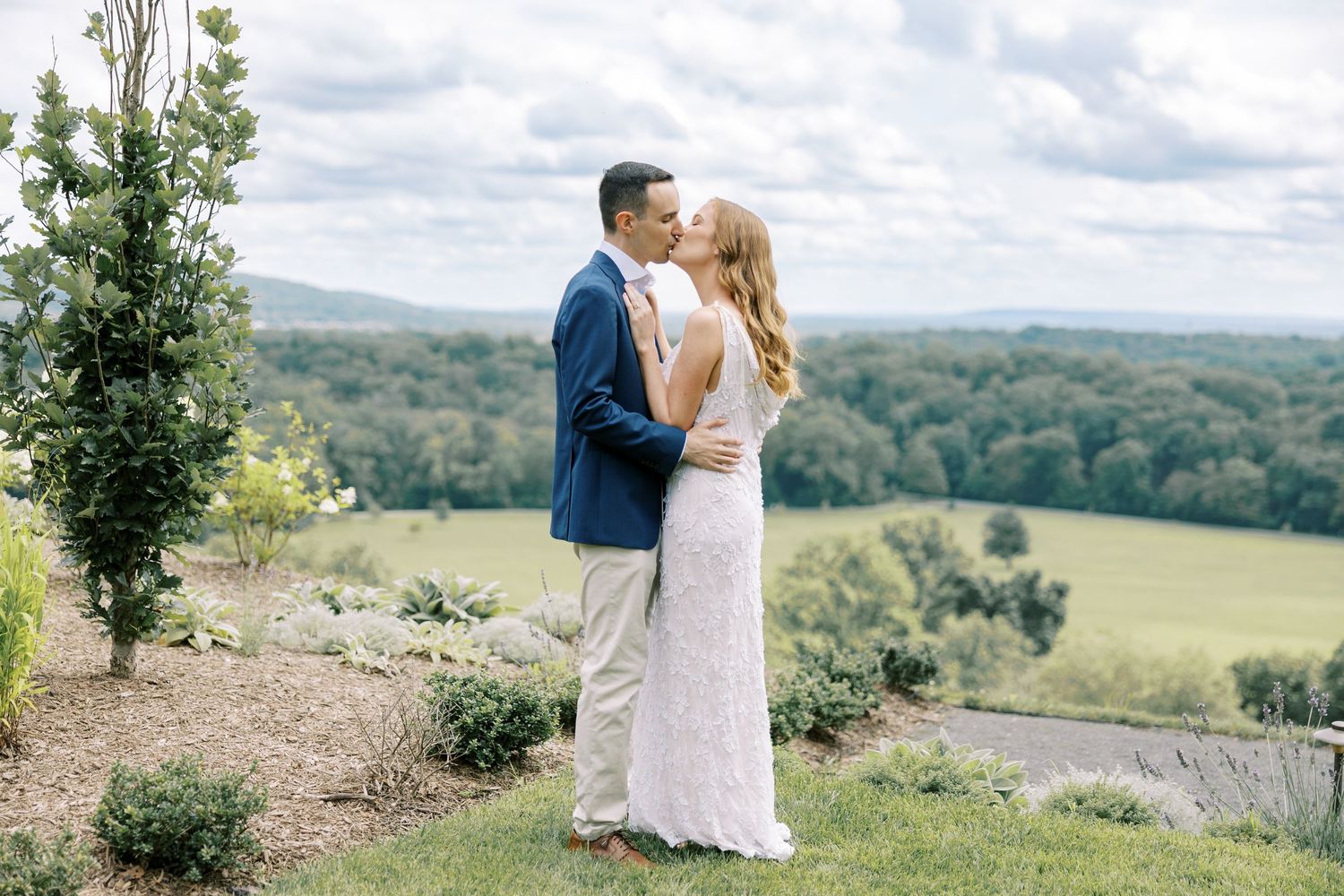 A couple in wedding attire shares an intimate moment at a scenic overlook with rolling hills and forests in the background at Pendry Natirar