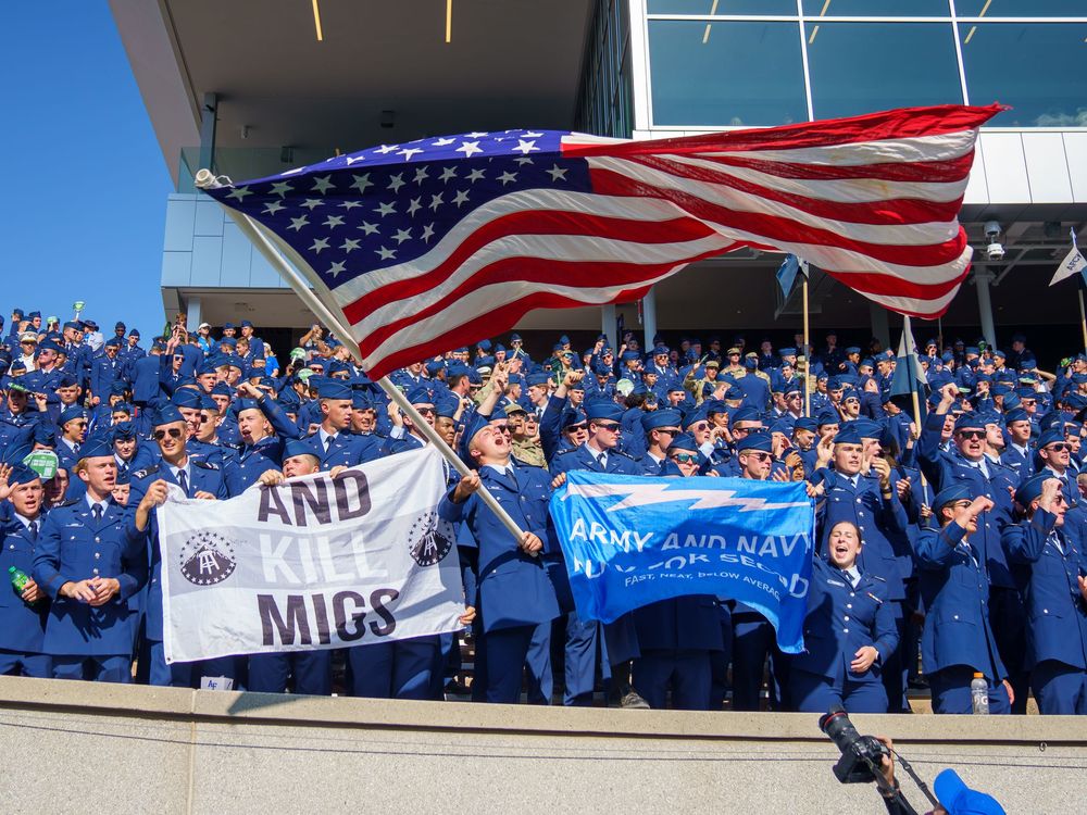 Large crowd waves American flag while cheering in stadium bleachers.