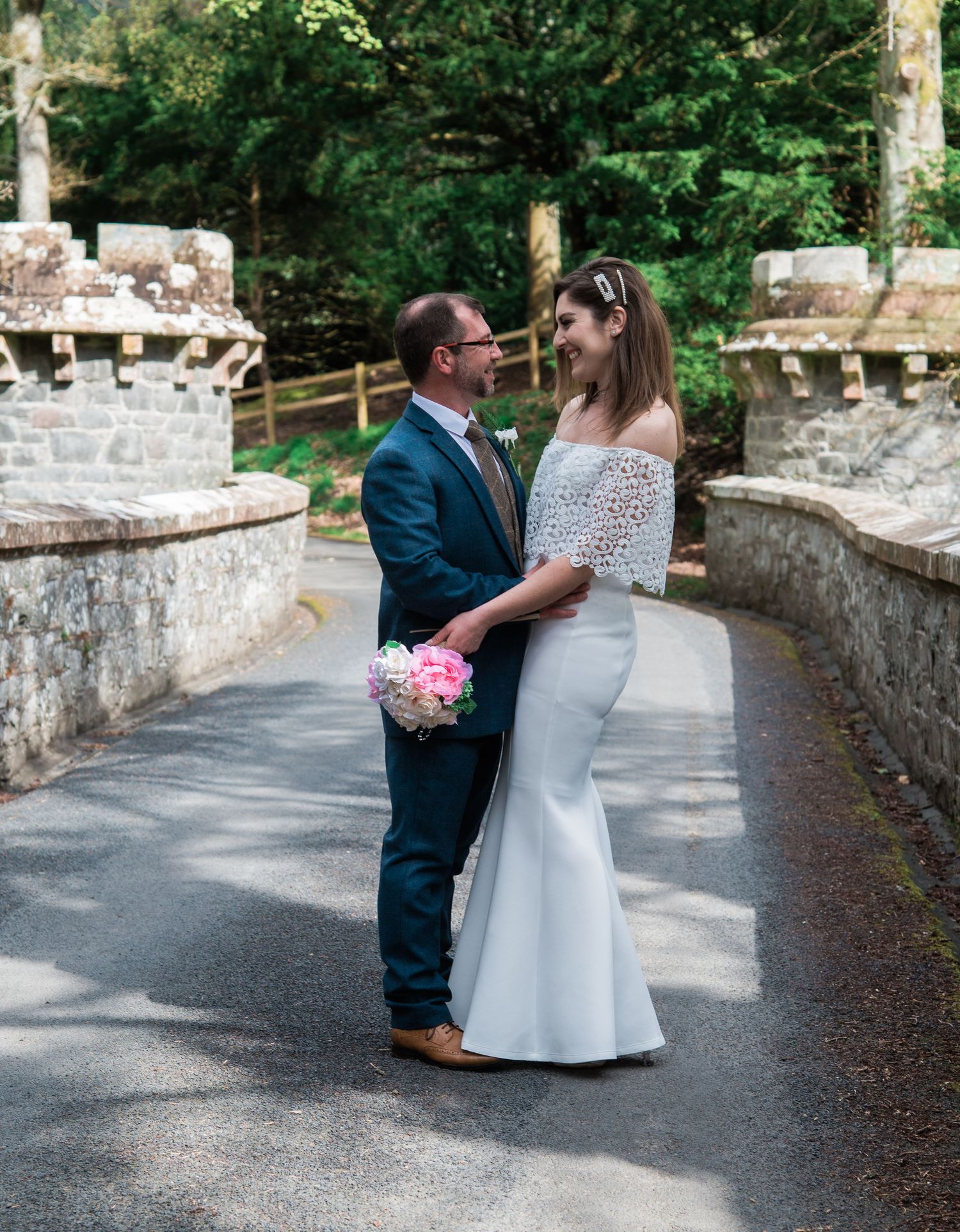 A formal couple embraces on a stone bridge path surrounded by lush greenery.