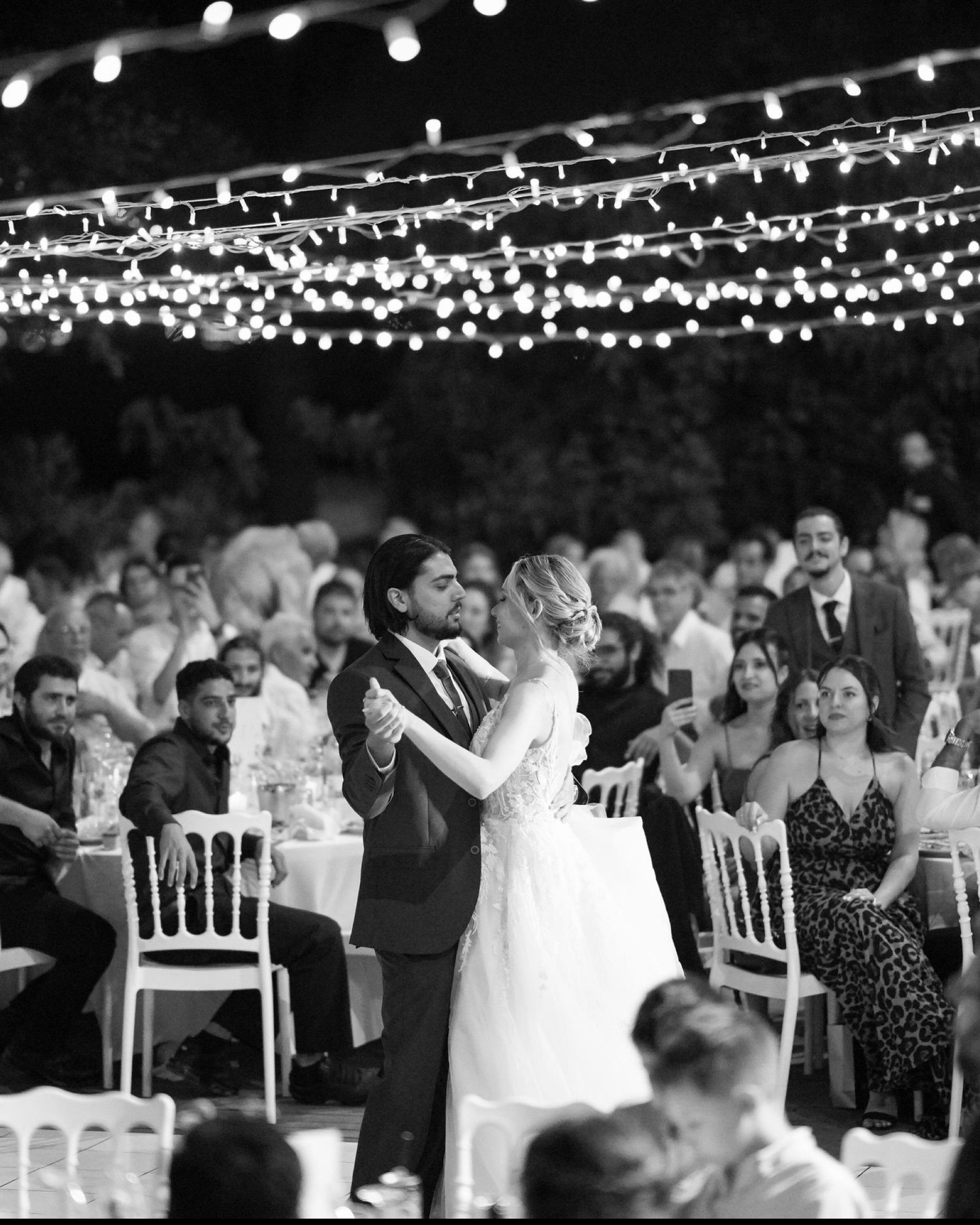 Black and white photo of a couple dancing at their wedding reception under romantic string lights with guests seated around.