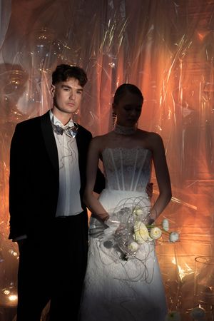 A couple poses in formal attire at a prom or wedding with dramatic orange lighting and shimmery backdrop.