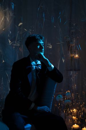A silhouette in a dark room lit by candles and blue light adjusts their bow tie in a moody atmospheric scene.