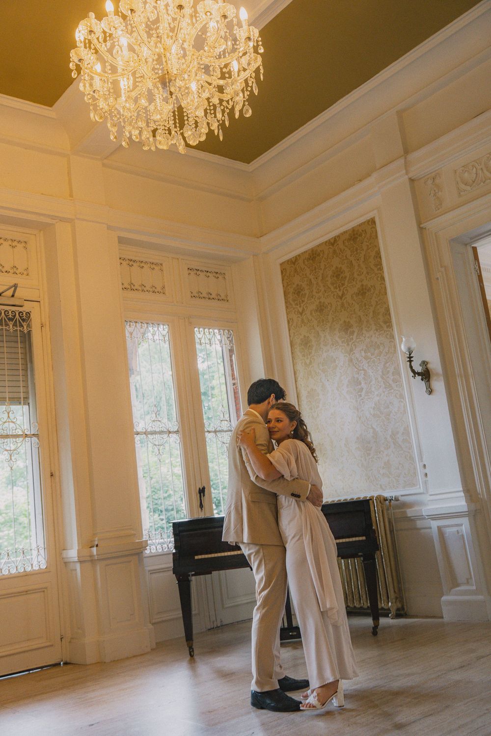 A couple embraces in an elegant room with a grand chandelier, piano, and ornate damask wallpaper.