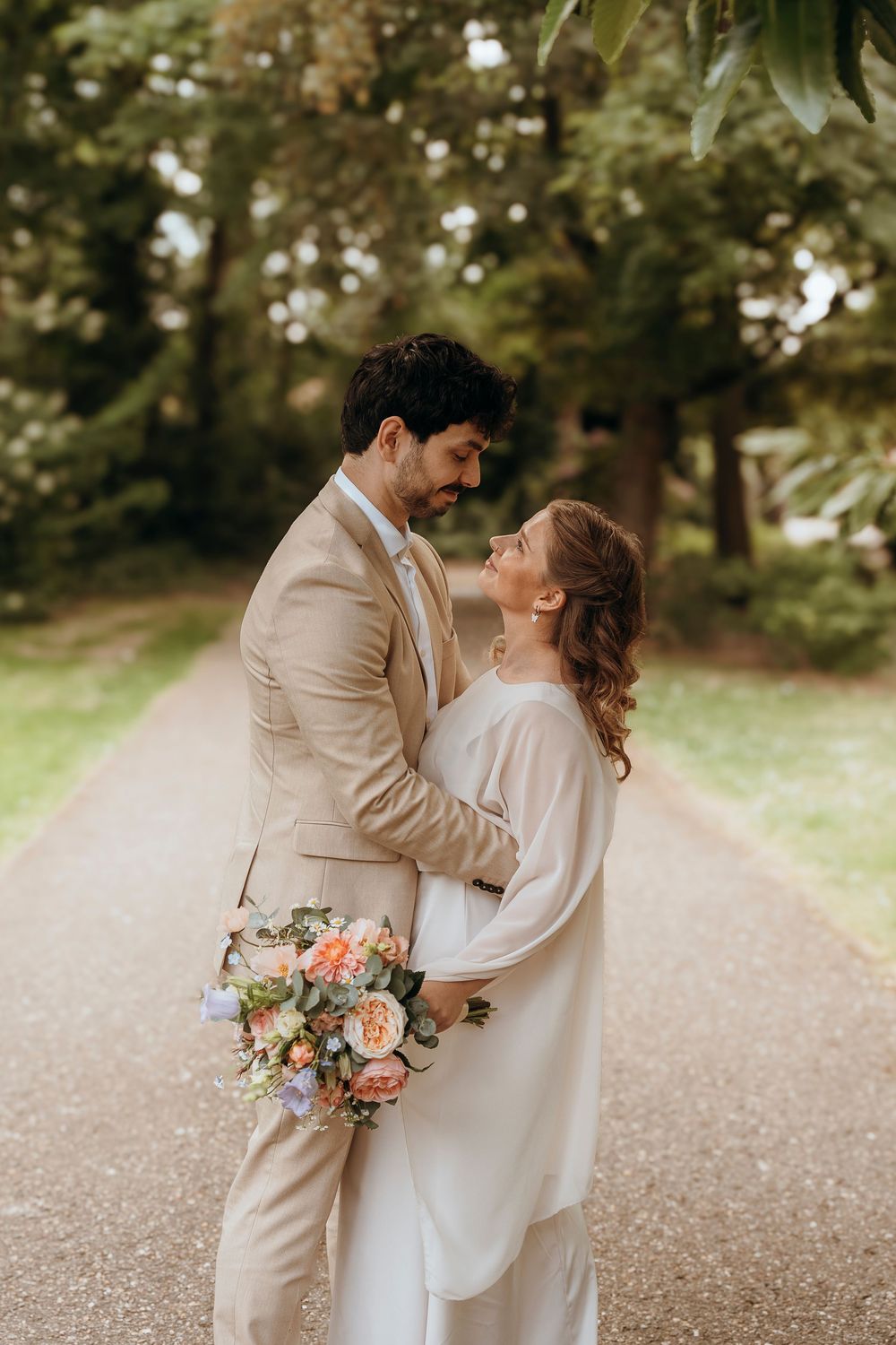 A romantic wedding couple embraces on a tree-lined path while holding a peachy pink floral bouquet.