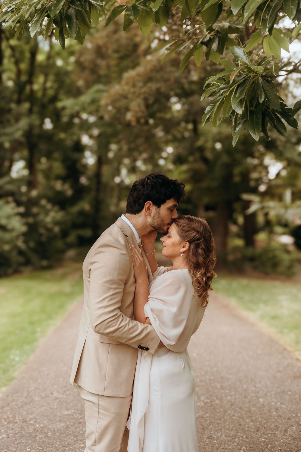 Two people embrace lovingly on a tree-lined path while wearing light colored formal attire.