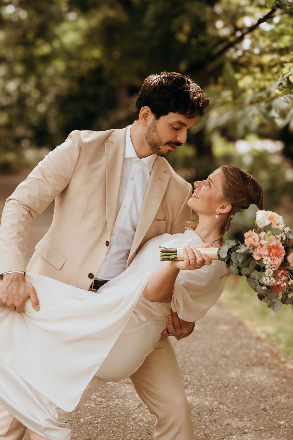 A romantic couple in matching beige attire dips and dances on a garden path while holding a peachy floral bouquet.