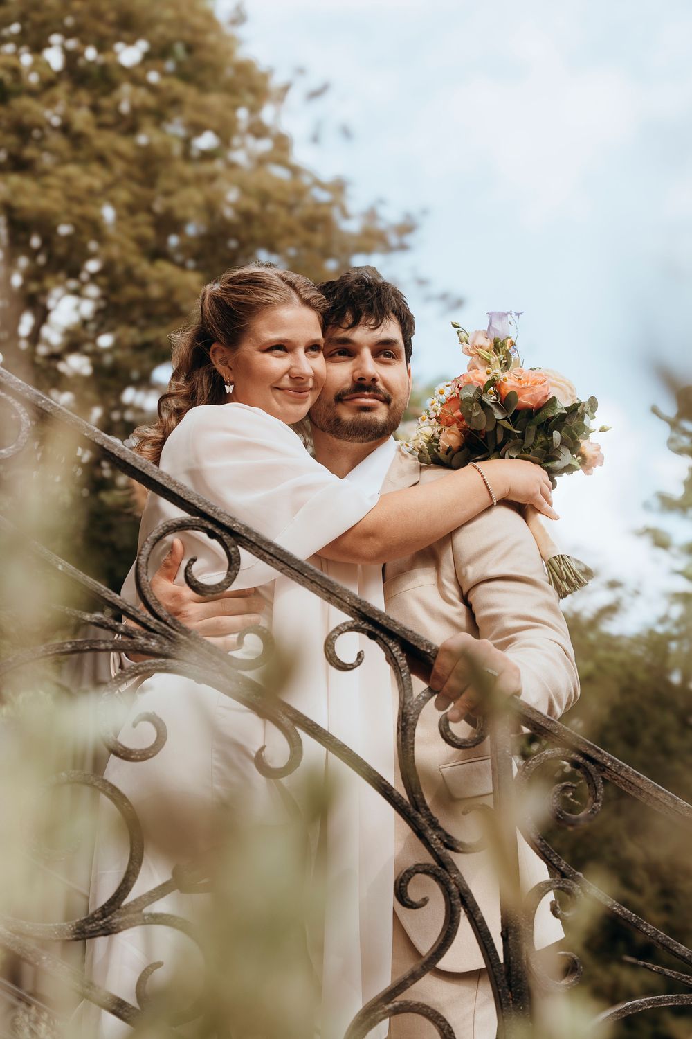 A romantic wedding couple in white outfits embrace on an ornate metal staircase while holding a peach-colored bouquet.