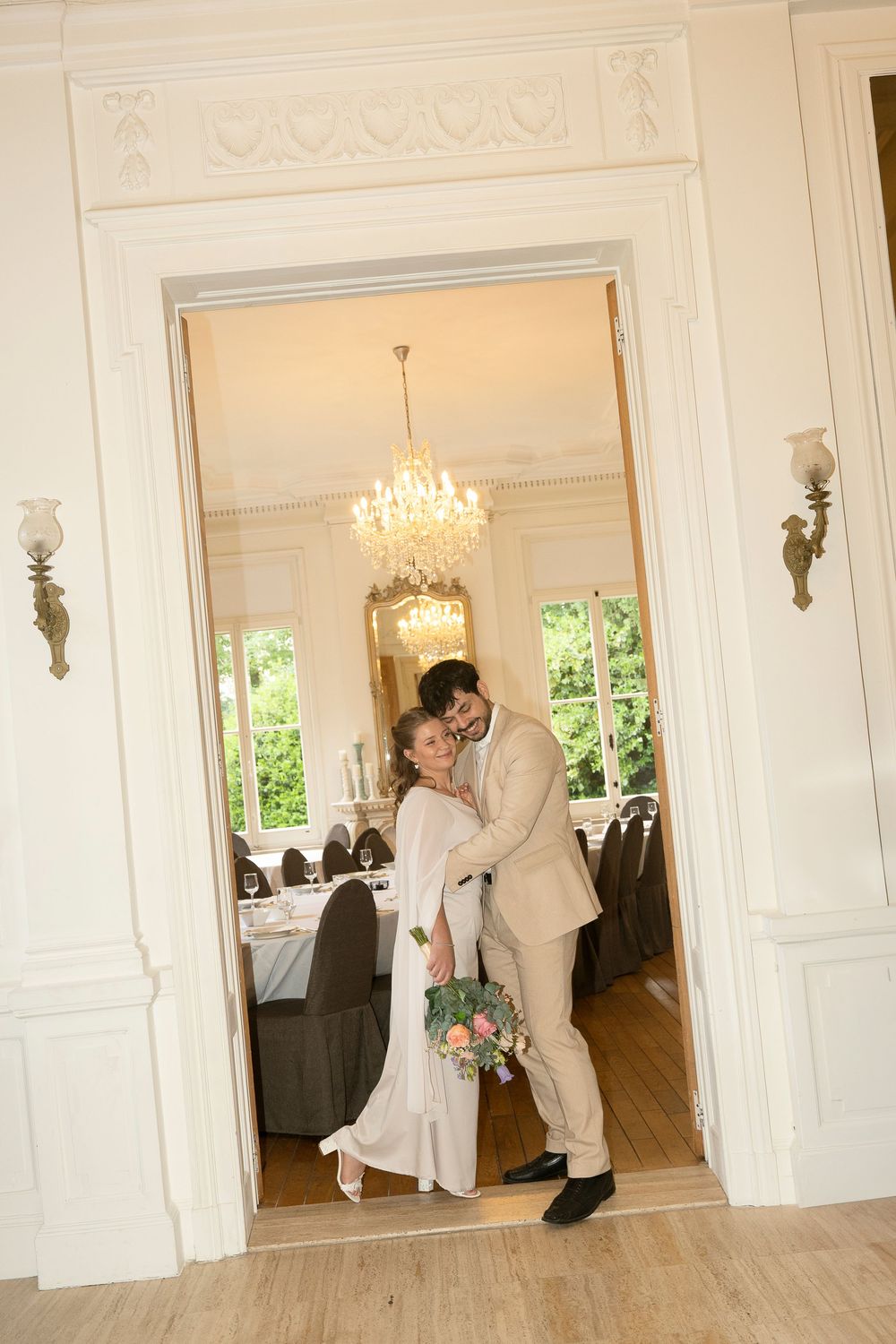 A couple shares an intimate dance in an elegant room with a crystal chandelier and ornate white moldings.