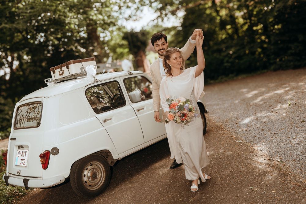 A newlywed couple dances near a vintage white Renault 4 car on a dirt path during golden hour.