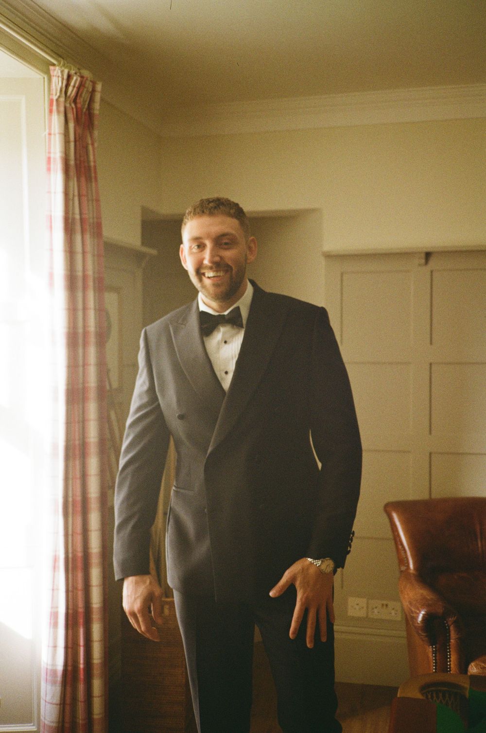 A groom wearing a black tuxedo and bow tie poses in a warmly lit room with curtained windows.