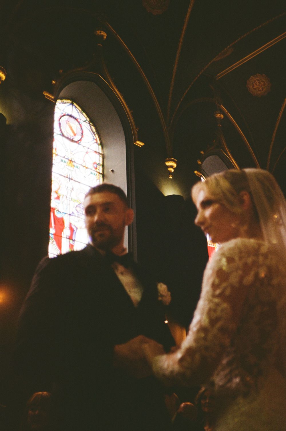 Two people in period costume converse in a dark room with stained glass windows visible in the background.