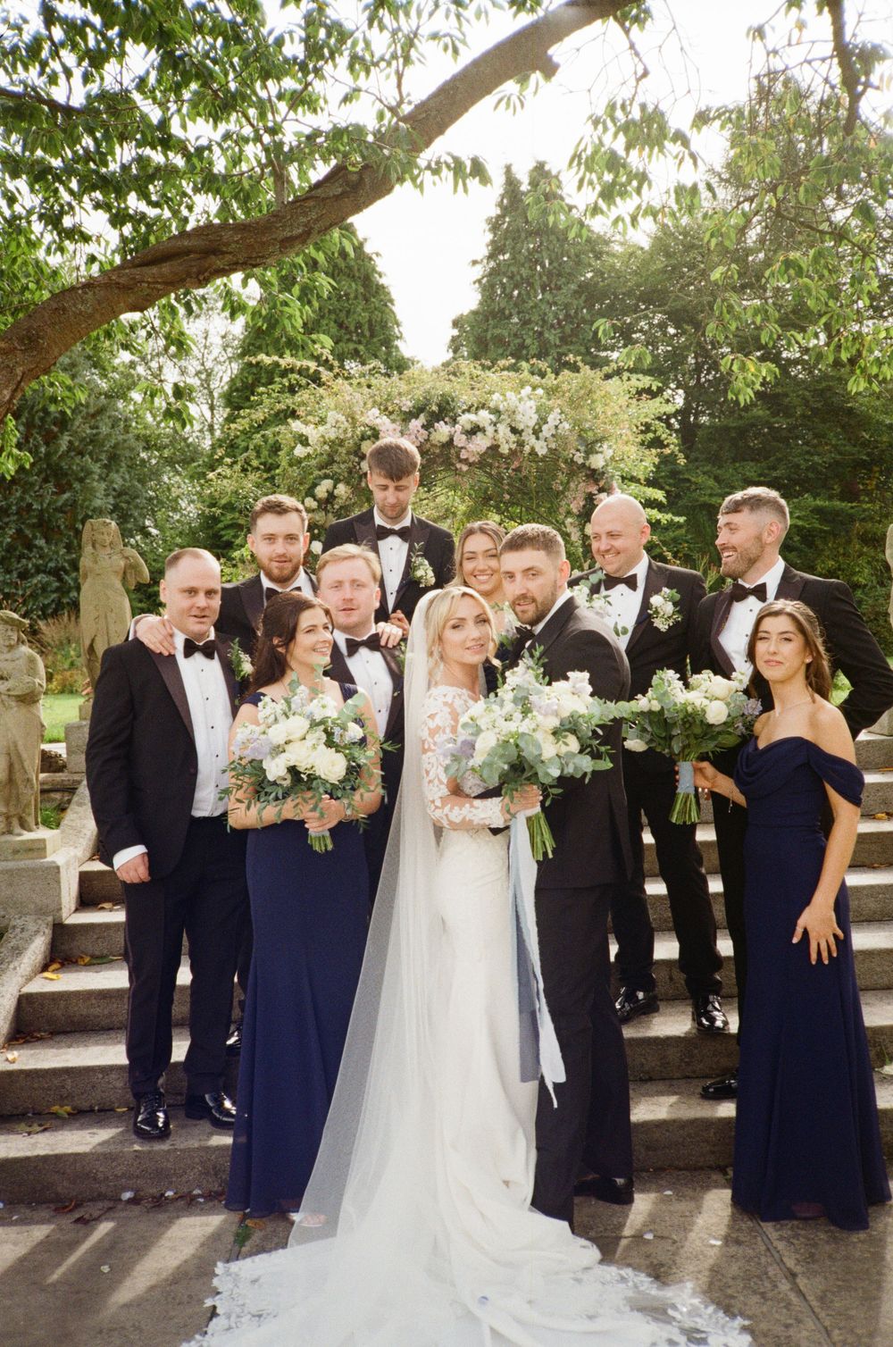 Wedding party in navy blue attire poses outdoors on stone patio with white floral bouquets.