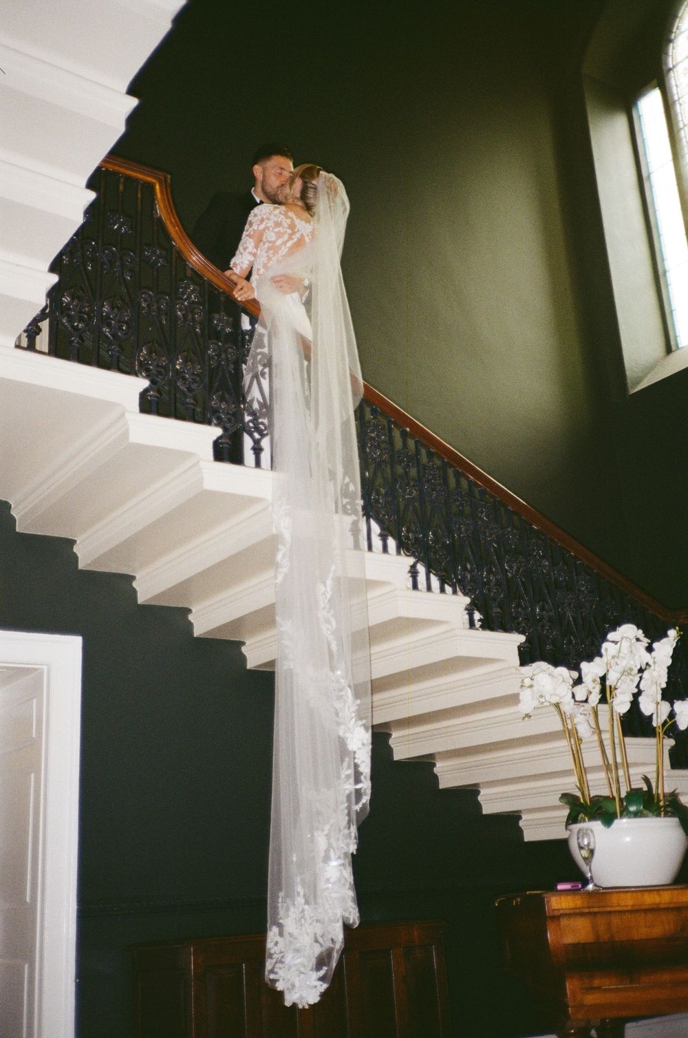 A bride in a flowing white lace wedding gown and veil stands gracefully on an elegant white staircase with dark walls.